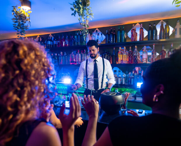 Young bartender preparing cocktails for cheering customers in a trendy bar with blue lights and hanging plants