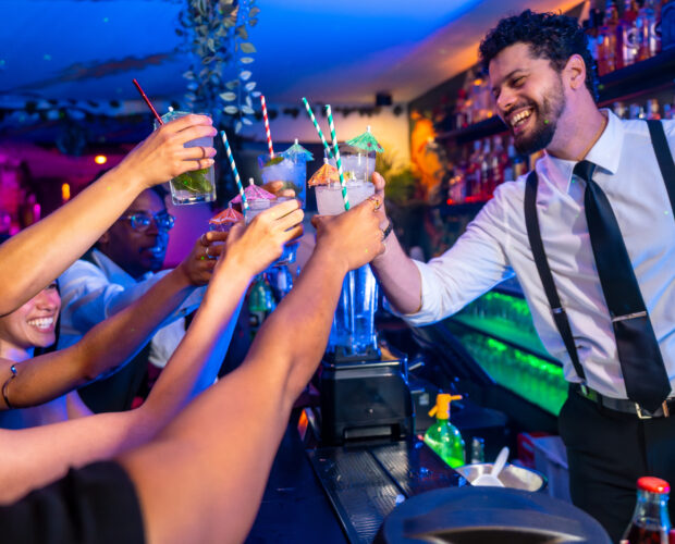 Happy friends raising cocktail glasses at nightclub bar counter, enjoying nightlife with bartender serving drinks
